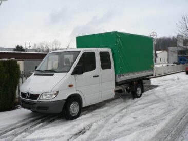 Weißer Mercedes-Benz Transporter mit einer grünen Plane auf der Ladefläche, geparkt auf schneebedeckter Straße.