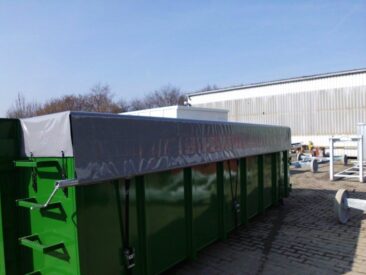 Grüner Abfallcontainer mit einer grauen Abdeckung auf der Oberseite, auf einem gepflasterten Boden.