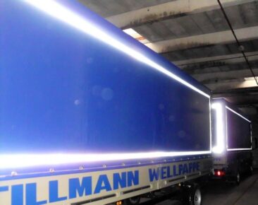 Zwei Lkw mit blauen Planen und einer hellen Beleuchtungslinie an den Seiten in einer Lagerhalle.