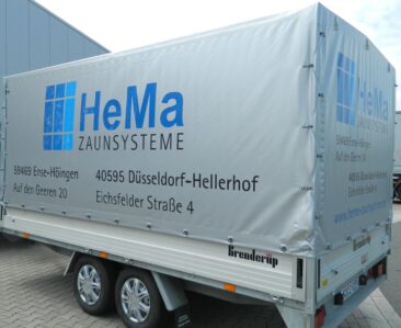 Ein Anhänger mit einer grauen Plane, die das Logo von HeMa Zaunsysteme zeigt.